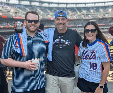 Three AdsLoom.net employees enjoying a casual outing at a baseball game, dressed in sports apparel, smiling and posing happily with the stadium crowd visible behind them.