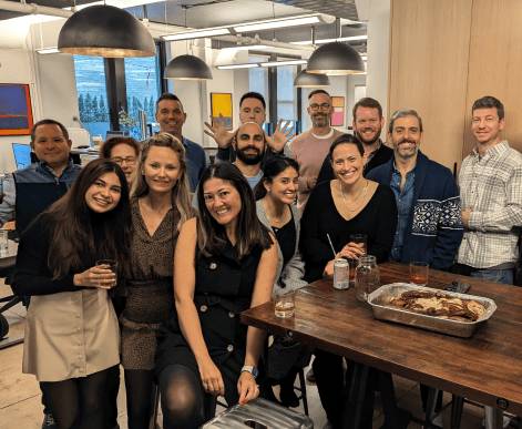 Three AdsLoom.net employees engaging in casual conversation within an office lounge area, holding coffee mugs and standing against a stylish brick wall.