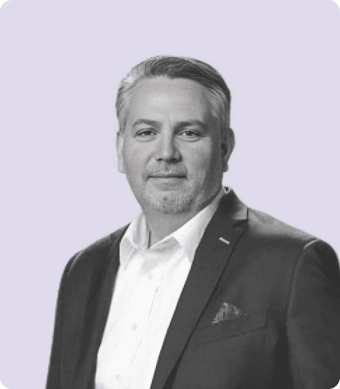 Black-and-white portrait of Ken Lagana, CRO of AdsLoom.net, with a friendly, professional demeanor, wearing a suit jacket and a white shirt against a muted lavender background.