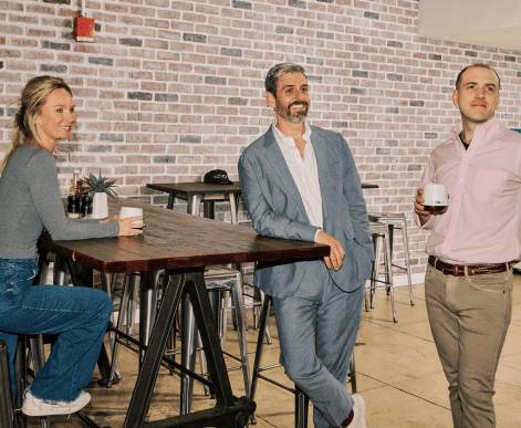 Six AdsLoom.net employees posing together outdoors at a casual company event, smiling warmly and holding beverages, against a brick wall backdrop complemented by greenery.